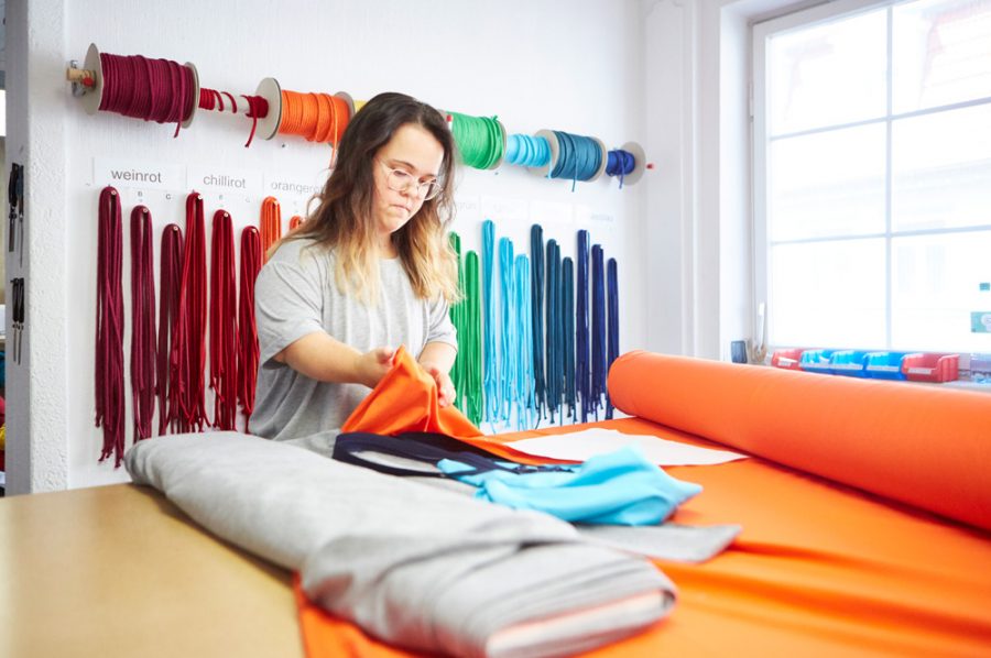 Nadine lässt am Schneidetisch ein orangefarbenes Stück Stoff durch die Finger gleiten. An der Wand hinter ihr hängen Spulen mit Kordeln in allen Farben, darunter kürzere Bänder an Nägeln.