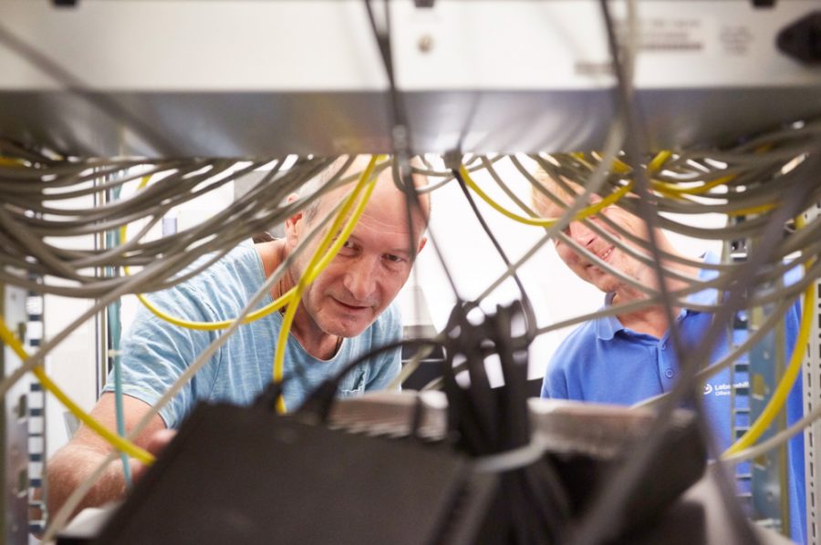 Blick durch die Verkabelung des Serverschrankes auf die lächelnden Gesichter von Michael und Nico.