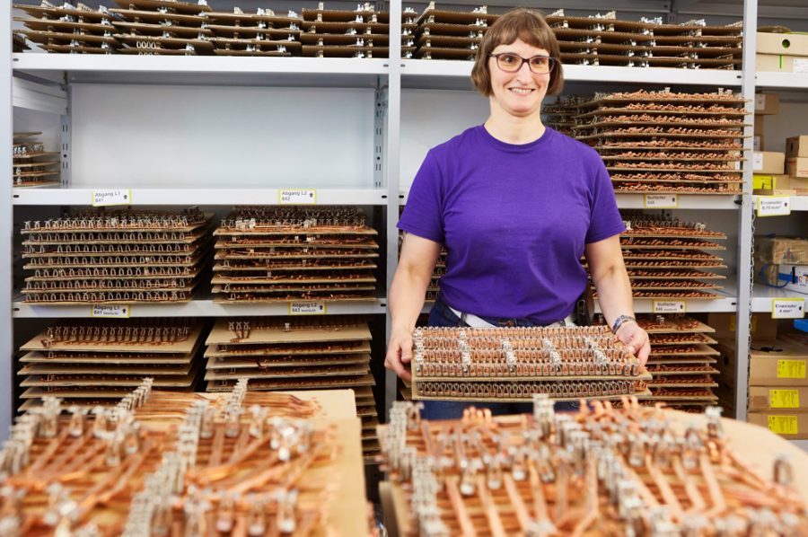 Hella hält einen Stapel mit Platten vor sich, auf denen kupferfarbene, längliche Metallstücke aufgereiht sind. Die Regale im Hintergrund sind gefüllt mit ebensolchen Platten.