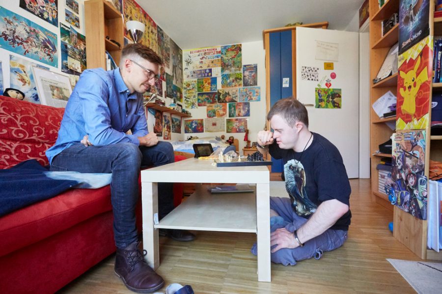 Florian und sein Begleiter von der Lebenshilfe beim Schachspiel. Der Begleiter sitzt auf dem roten Sofa, Florian auf dem Boden vor dem Couchtisch.