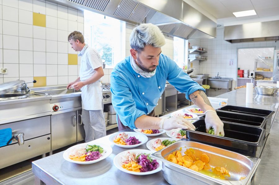 Auf der Arbeitsfläche richtet Guiliano bunte Salatteller an. Im Hintergrund spritzt Nico das Spülbecken aus.