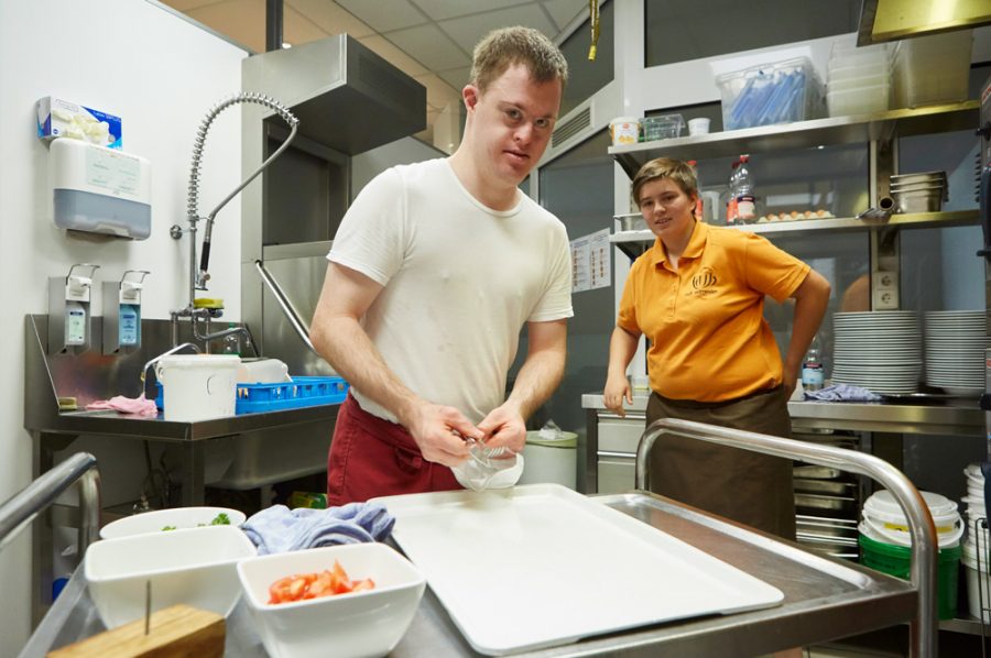 Florian und Tamara in der Küche. Florian steht hinter einem Edelstahl-Servierwagen, Tamara lehnt im Hintergrund an einer Arbeitsfläche mit Tellerstapeln.