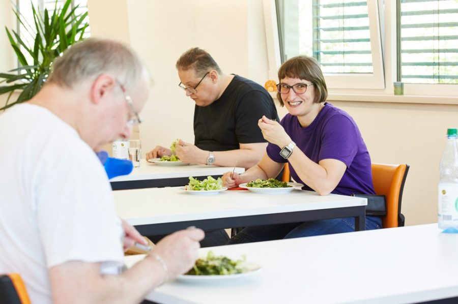 Hella und zwei Kollegen an getrennten Tischen beim Mittagessen. Die Tische stehen in großem Corona-Abstand zueinander. Links im Vordergrund sitzt der ältere Kollege, mit dem Hella sich besonders gut versteht.