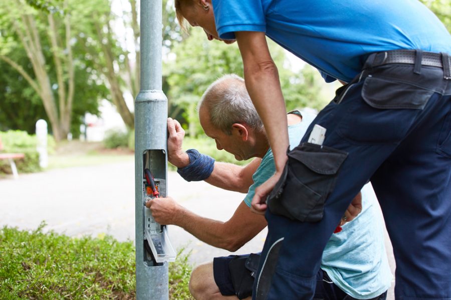 Vor dem geöffneten Sicherungskasten im Sockel einer Straßenlaterne kniet Michael, Nico schaut ihm über die Schulter.