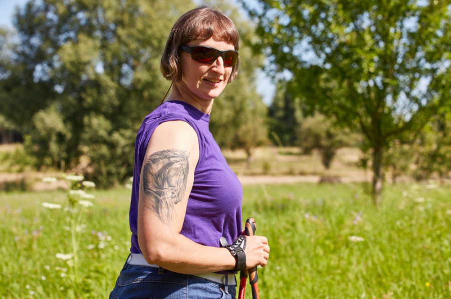 Hella mit Walking-Stöcken und Sonnenbrille vor einer grünen Wiese. Auf dem rechten Oberarm sieht man das äußerst fein gestochene Tattoo eines Pferdekopfes.