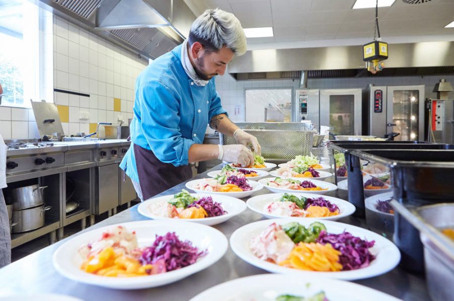 Guiliano rundet eine Salatmischung in einer langen Reihe von beinahe fertig angerichteten Salattellern ab. Aus dem Geschirr leuchtet es lila, gelb, grün und rosé.