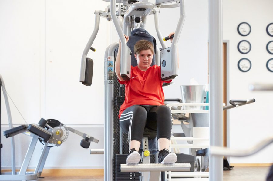 Tamara im Fitnessstudio. Sie sitzt auf einem grau und schwarz gehaltenen Hightech-Trainingsgerät, bei dem zwei Platten mit den Armen gegeneinanderzudrücken sind. Ihr Gesicht verrät die Anstrengung.