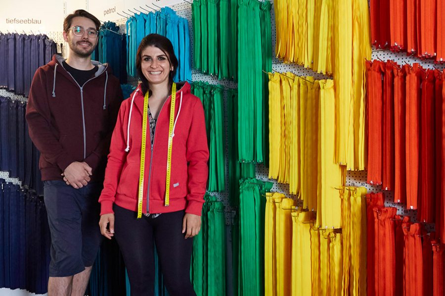 Die strahlende Yaprak steht mit dem Kollegen Leon vor frei hängenden, langen Reißverschlüssen in allen Farben des Regenbogens.